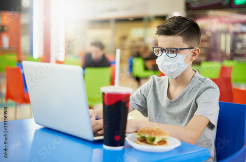 Teenager wearing glasses and a protective mask uses a laptop in a restaurant before lunch
