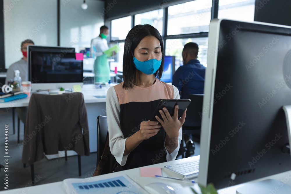 Asian woman wearing face mask using digital tablet while sitting on her desk at modern office Asian woman wearing face mask using digital tablet while sitting on her desk at modern office