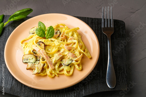 beige plate of pasta with ham and mushrooms in a creamy sauce with basil on a black board with a fork.