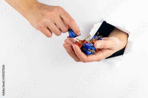 Female hands are holding gifts on a white background. A hand goes through a hole in a white paper wall and holds gifts. The other hand takes one gift.