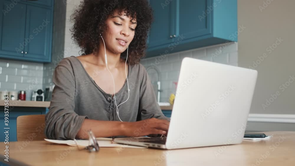 Smiling happy african woman sitting indoors at home and using laptop computer while listening music with earphones