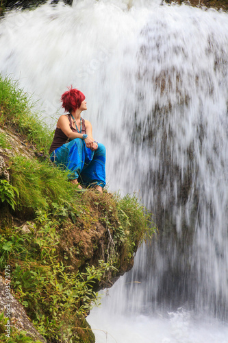a young woman with red hair dressed in boho style sits in the background of a waterfall