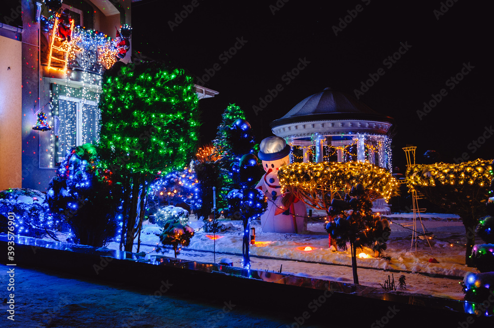 Fototapeta House Adorned with Christmas Holiday Lights and Decorations including Santa Snowman and Giant Trees Illuminated at Night. Christmas Lights outside on a House