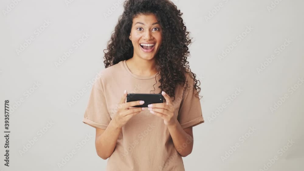 A surprised african american woman is using her smartphone holding it horizontally standing isolated over beige background A surprised african american woman is using her smartphone holding it horizontally standing isolated over beige background