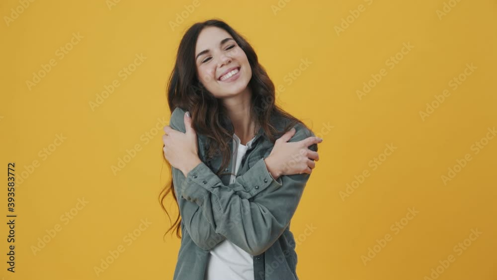 A smiling woman is hugging herself standing isolated over yellow background A smiling woman is hugging herself standing isolated over yellow background