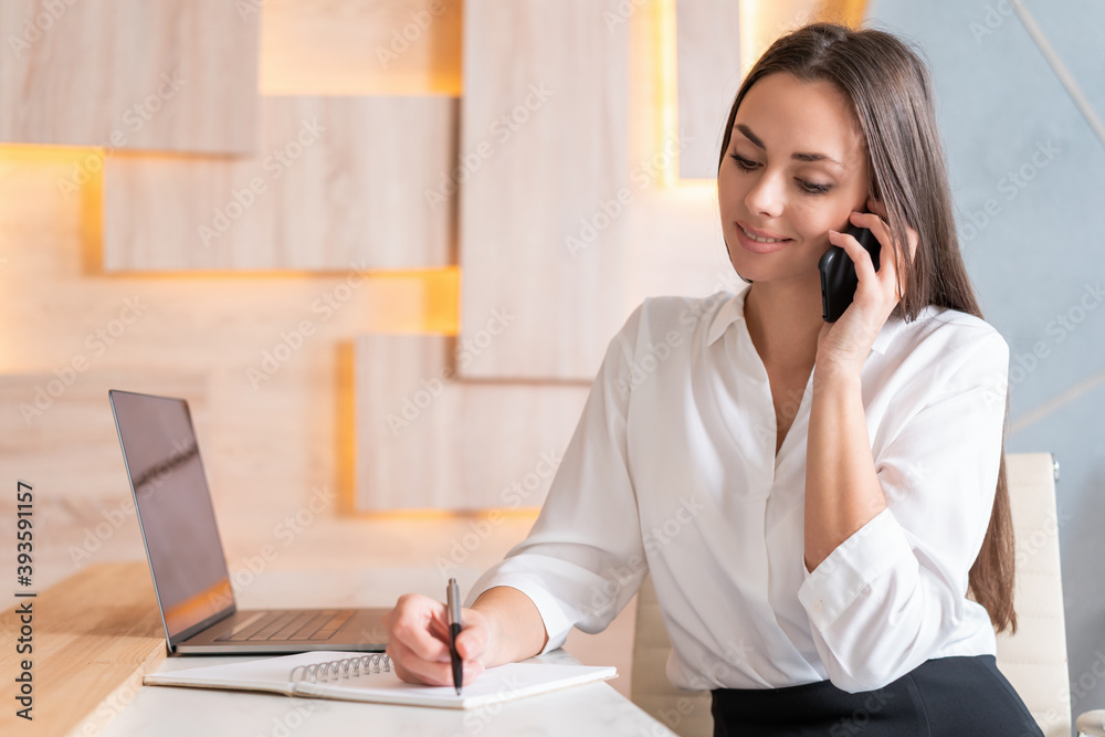 Woman office manager with phone in hand, smiling making a note in notebook sitting to office desk. Middle shot of smiling friendly secretary taking a call. Concept of secretary Woman office manager with phone in hand, smiling making a note in notebook sitting to office desk. Middle shot of smiling friendly secretary taking a call. Concept of secretary
