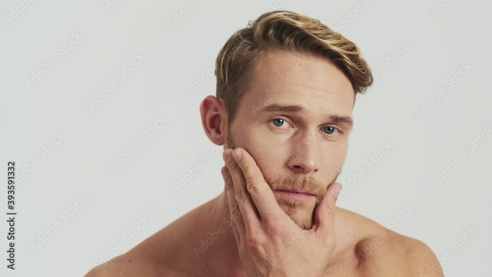 A positive half-naked blonde man is touching his beard while looking to the camera like to the mirror standing isolated over a grey background in the studio