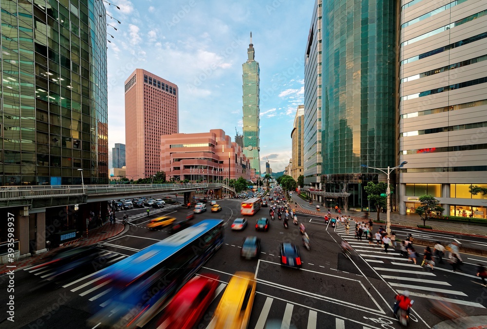 Fototapeta premium View of a busy street corner in Downtown Taipei City at rush hour with cars & buses dashing by, Taipei 101 Tower & World Trade Center Building in Xinyi Financial District & people passing on crosswalk