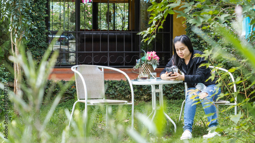Asian woman wears a gray shirt and white jeans sitting in the garden in front of the house, holding a smartphone and a black coffee cup on the table.