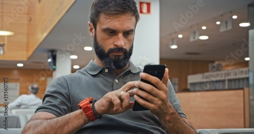 Handsome bearded man texting message on mobile phone while sitting at hospital lobby waiting doctor visit. Young man using virtual assistant app to sets reminder