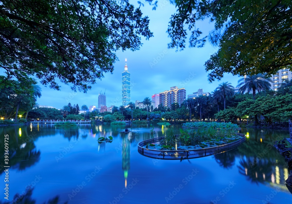 Lakeside scenery of Taipei 101 Tower among skyscrapers in Xinyi District Downtown at night with view of reflections on the pond in an urban park ~ Romantic nightscape of Taipei city under gloomy sky