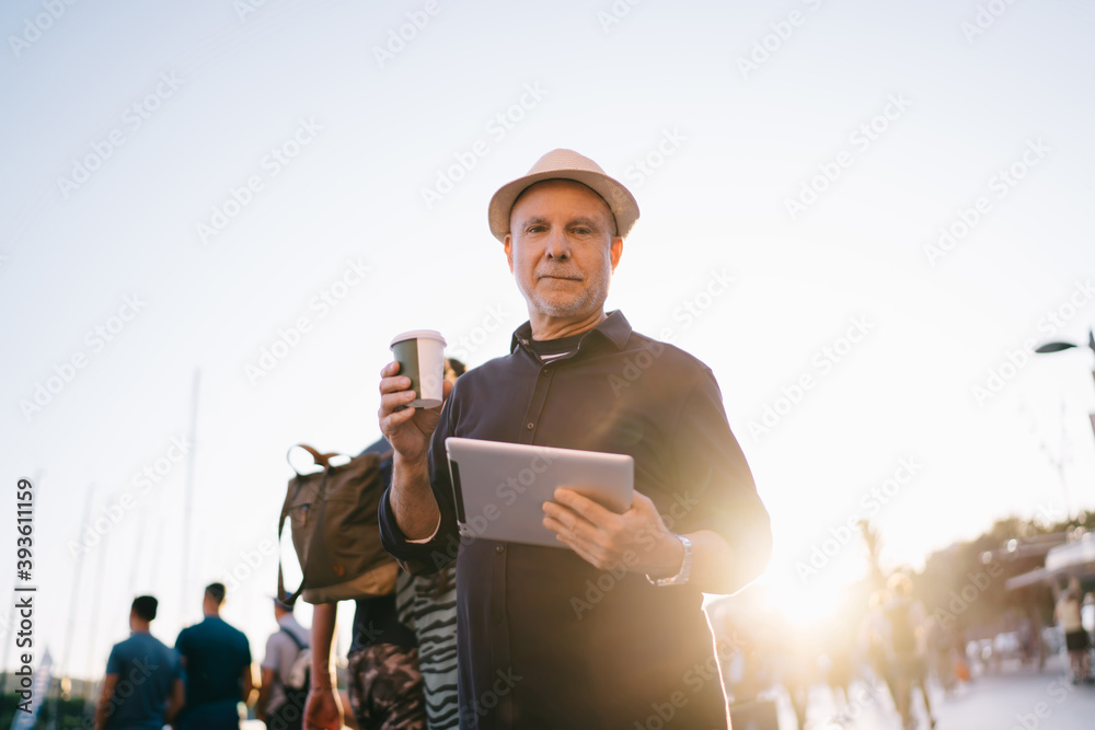 Half length portrait of good looking male tourist with takeaway caffeine beverage and touch pad posing at city street, backlit of Caucasian man on retirement spending sunny daytime for networking Half length portrait of good looking male tourist with takeaway caffeine beverage and touch pad posing at city street, backlit of Caucasian man on retirement spending sunny daytime for networking