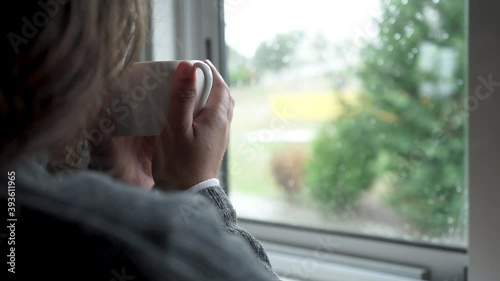 Camera panning from the back of a woman drinking tea or coffee inside the house looking through the window while it's raining outside.