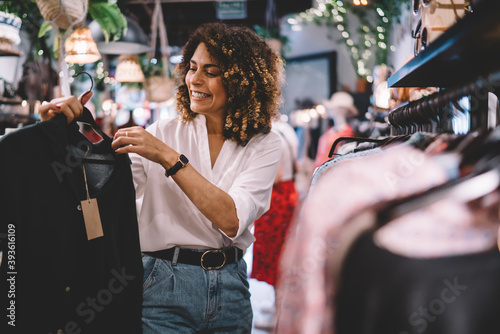Cheerful female standing near hangers with brand clothes and laughing during Black Friday shopping with discounts, happy hipster girl enjoying pastime for update her wardrobe spending day in boutique photo