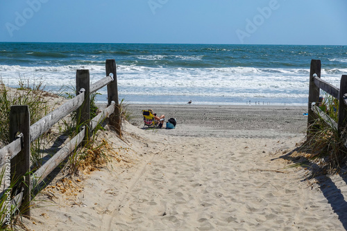 View along a sand path with a wooden fence towards an empty beach with one girl sunbather siting on a chair