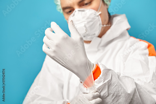Close-up of doctor in protective clothing putting on sterile glove over plain blue background. Environmental friendliness and safety during medical procedures.