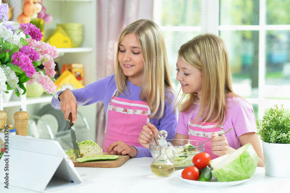 Two girls in pink aprons preparing salad on kitchen table with tablet Two girls in pink aprons preparing salad on kitchen table with tablet