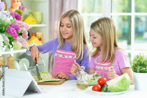 Two girls in pink aprons preparing salad on kitchen table with tablet