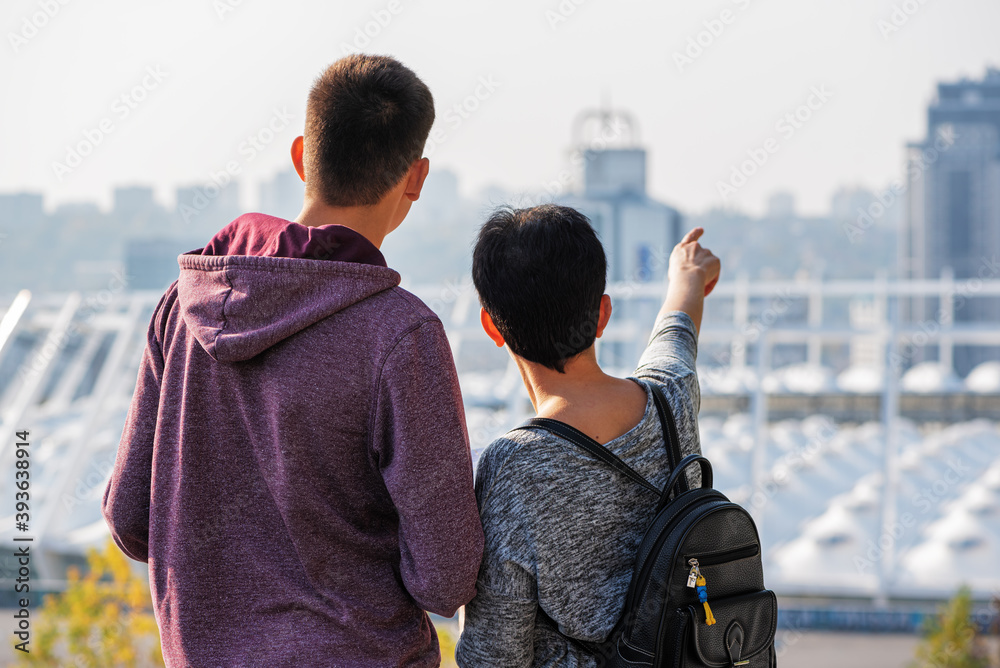 Two young caucasian people tourists exploring new city together. Girl with backpack pointing her finger at the building. Back view.