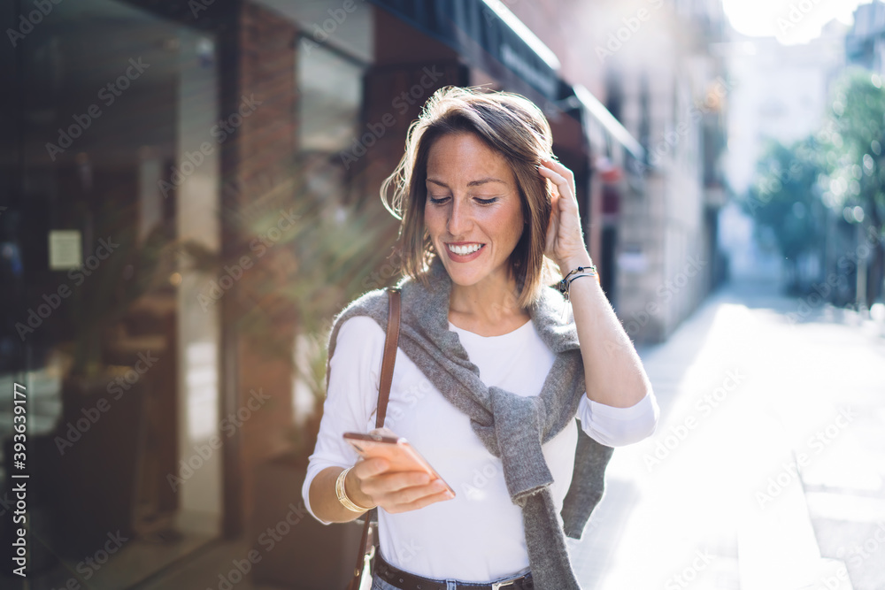 Cheerful pretty caucasian woman walking on city street making payment via banking app on mobile phone, smiling female influencer chatting and sending messages on smartphone strolling in town