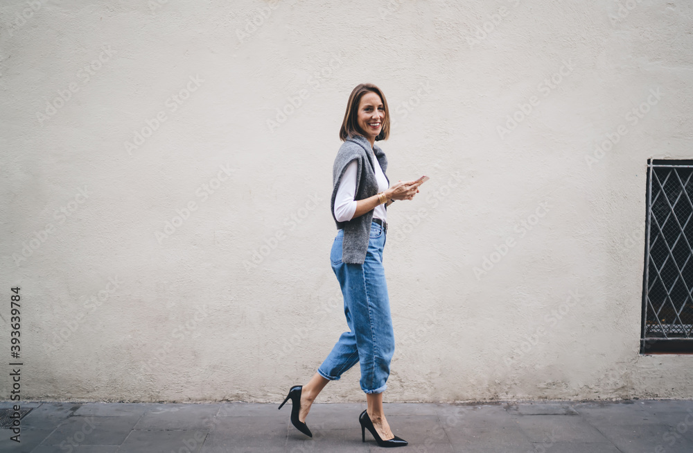 Side full length portrait of cheerful Caucasian woman dressed in stylish streetwear holding modern cellphone gadget and smiling at camera while walking near urban wall, happy female blogger laughing Side full length portrait of cheerful Caucasian woman dressed in stylish streetwear holding modern cellphone gadget and smiling at camera while walking near urban wall, happy female blogger laughing
