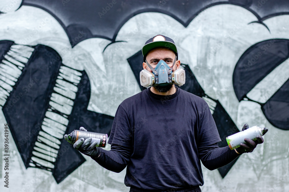 Graffiti artist posing in front of his drawing on the wall, with two  aerosol spray paints in a can, wearing protective face mask / respirator  with filters. Street art culture concept. Stock