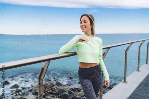 Cheerful sportswoman dressed in comfortable tracksuit standing at seashore pier and smiling during workout break, happy Caucasian athlete enjoying leisure for training body muscles at coastline
