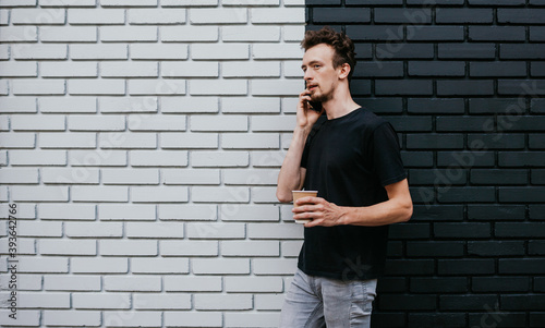 A young charismatic guy, a hipster, drinks coffee and speaks on the phone against the background of a brick wall. Dressed in a black T-shirt. Mock-up for printing