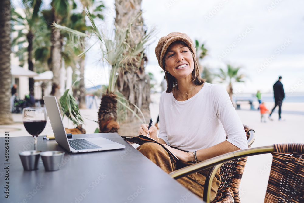 Positive caucasian female in trendy wear making notes in planner working remotely on cafe interior, creating young woman journalist working remotely making publication for blog use laptop computer