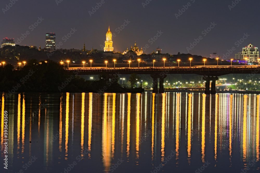 Night cityscape with Paton bridge over Dnieper river. Light from lanterns reflected in the water. Illuminated Bell tower of Kyiv Pechersk Lavra at the background. City lights reflected in the water