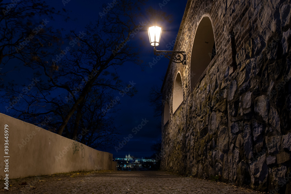 
light from street lighting and old stone fortress walls from the 15th century and paving blocks on the ground for pedestrians at night in the center of prague in the czech republic and in the backgro