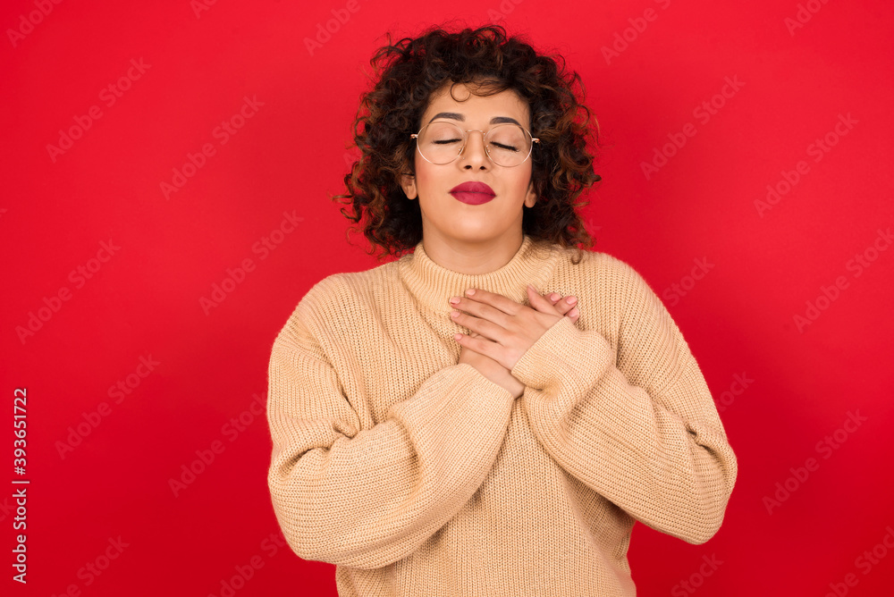 Young beautiful Arab woman wearing beige sweater closes eyes and keeps hands on chest near heart, expresses sincere emotions, being kind hearted and honest. Body language and real feelings concept. Young beautiful Arab woman wearing beige sweater closes eyes and keeps hands on chest near heart, expresses sincere emotions, being kind hearted and honest. Body language and real feelings concept.