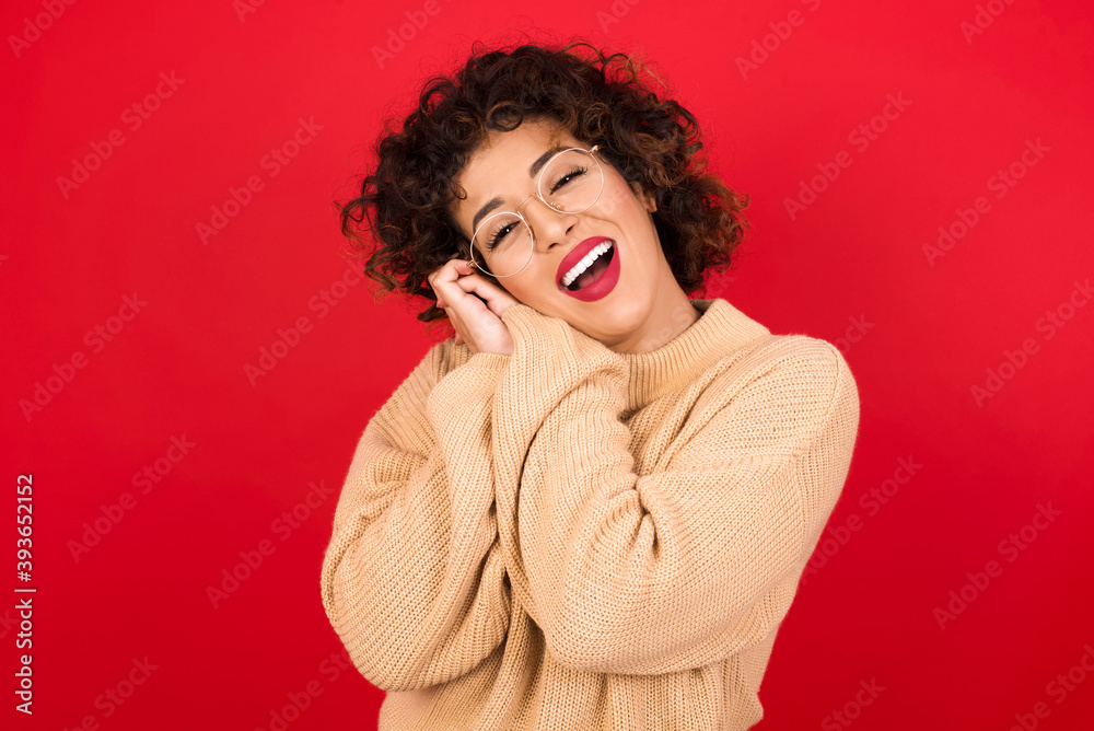 Dreamy Young beautiful Arab woman wearing beige sweater against red background with pleasant expression, closes eyes, keeps hands crossed near face, thinks about something pleasant Dreamy Young beautiful Arab woman wearing beige sweater against red background with pleasant expression, closes eyes, keeps hands crossed near face, thinks about something pleasant