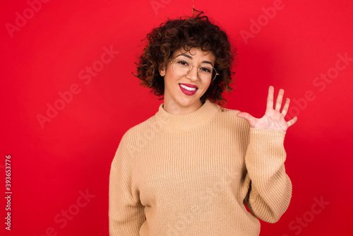 Young beautiful Arab woman wearing beige sweater against red background showing and pointing up with fingers number five while smiling confident and happy.