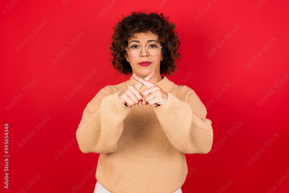 Young beautiful Arab woman wearing beige sweater against red background has rejection angry expression crossing fingers doing negative sign. Young beautiful Arab woman wearing beige sweater against red background has rejection angry expression crossing fingers doing negative sign.