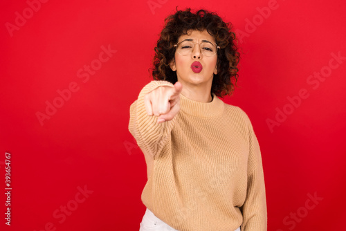 Cheerful Young beautiful Arab woman wearing beige sweater against red background indicates happily at you, chooses to compete, has positive expression, makes choice. © Jihan