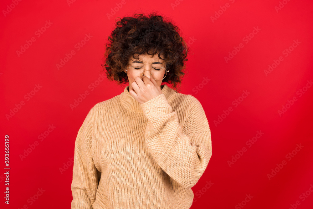 Young beautiful Arab woman wearing beige sweater against red background, holding his nose because of a bad smell.