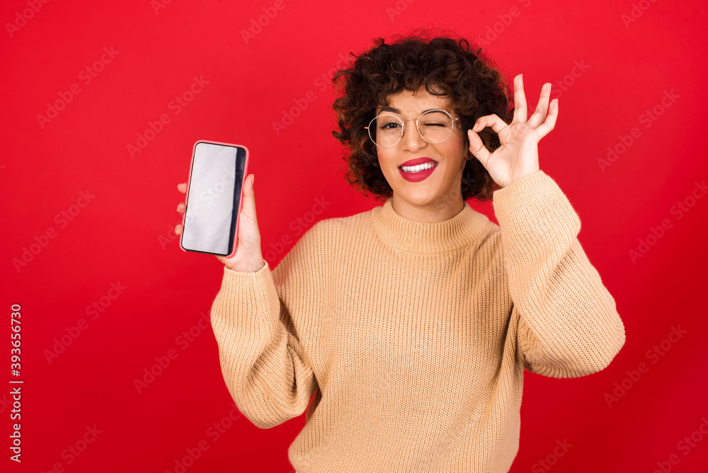 Excited Young beautiful Arab woman wearing beige sweater against red background showing smartphone blank screen, blinking eye and doing ok sign with hand.  Advertisement concept.