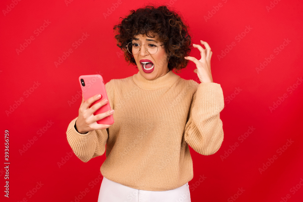 Photo of outraged annoyed Young beautiful Arab woman wearing beige sweater against red background holds cell phone, makes call, argues with colleague, expresses negative emotions. People and anger. Photo of outraged annoyed Young beautiful Arab woman wearing beige sweater against red background holds cell phone, makes call, argues with colleague, expresses negative emotions. People and anger.
