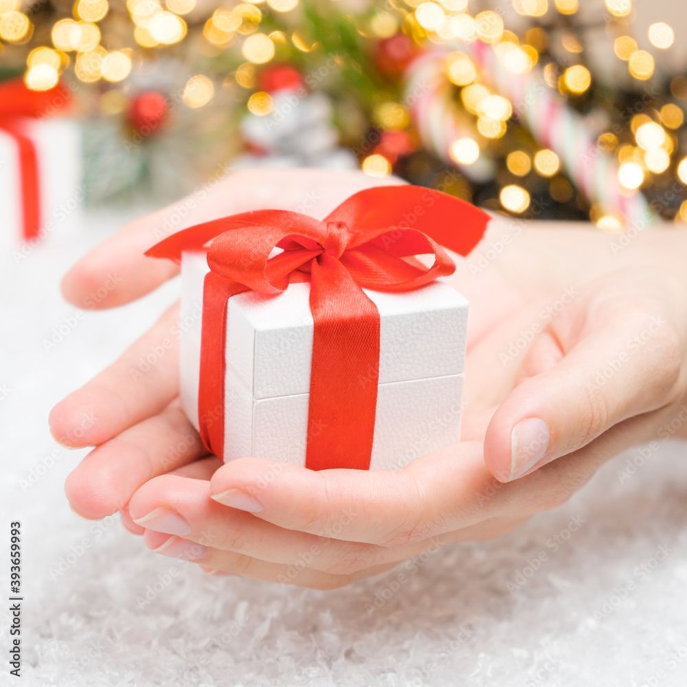 Close up woman hand holding a white gift box with a red ribbon on the background of presents, candy stick, Christmas tree and golden lights Close up woman hand holding a white gift box with a red ribbon on the background of presents, candy stick, Christmas tree and golden lights