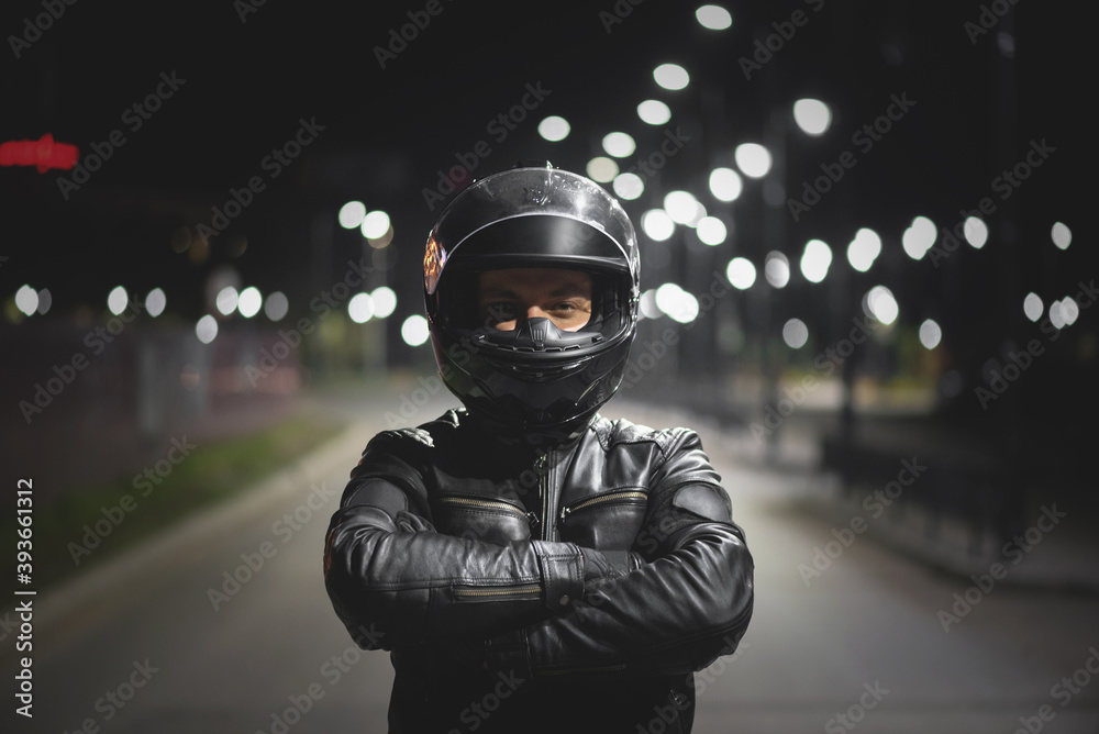 Motor biker in a helmet with open visor is standing on the night road.