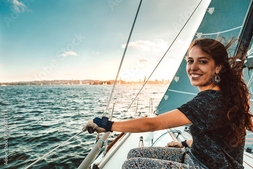 Beautiful young traveler woman smiling at the camera while navigating on a sailing boat. City of Sydney, Australia in the background. Concept about lifestyle, leisure, sport, travel and people. 