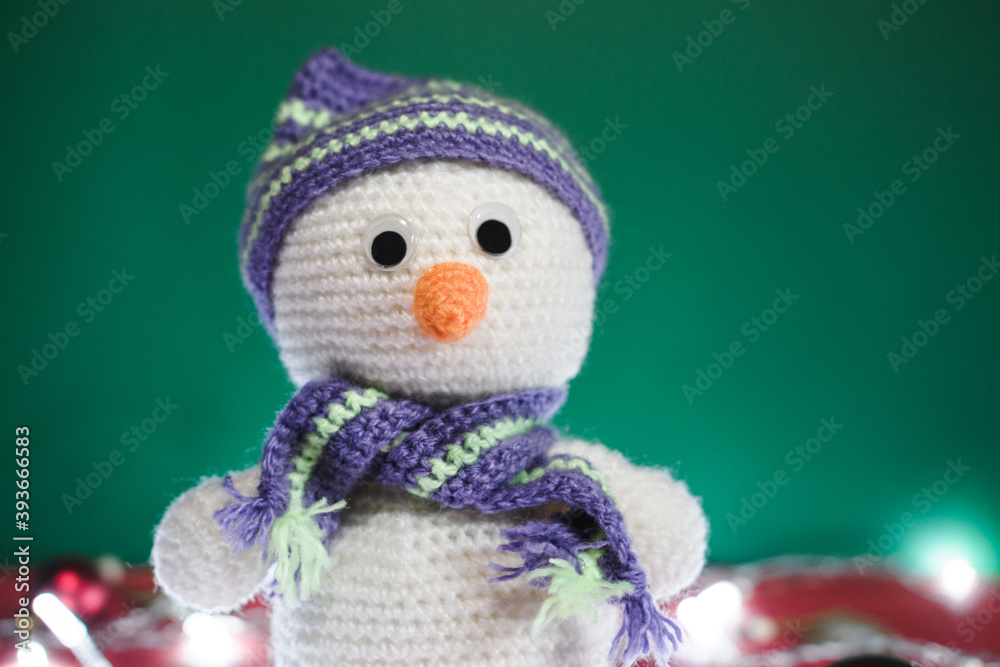 Cute knitted white snowman with a carrot for a nose and a striped hat on a green background, horizontal Christmas banner. New year's card with a handmade toy. Cute knitted white snowman with a carrot for a nose and a striped hat on a green background, horizontal Christmas banner. New year's card with a handmade toy.