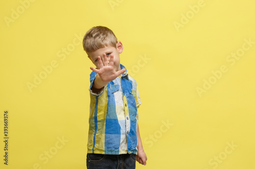 Little cute boy in a bright shirt put his hand palm forward in a prohibiting gesture isolated on a yellow background.
