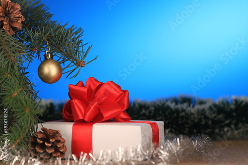 New year's gift under the branches of the tree. White gift box with red ribbon and red bow on a blue background. With space for text.