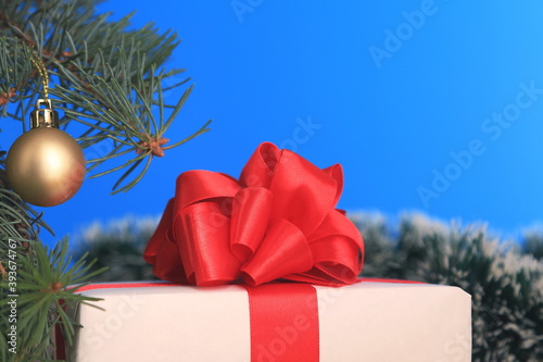 New year's gift under the branches of the tree. White gift box with red ribbon and red bow on a blue background. With space for text.