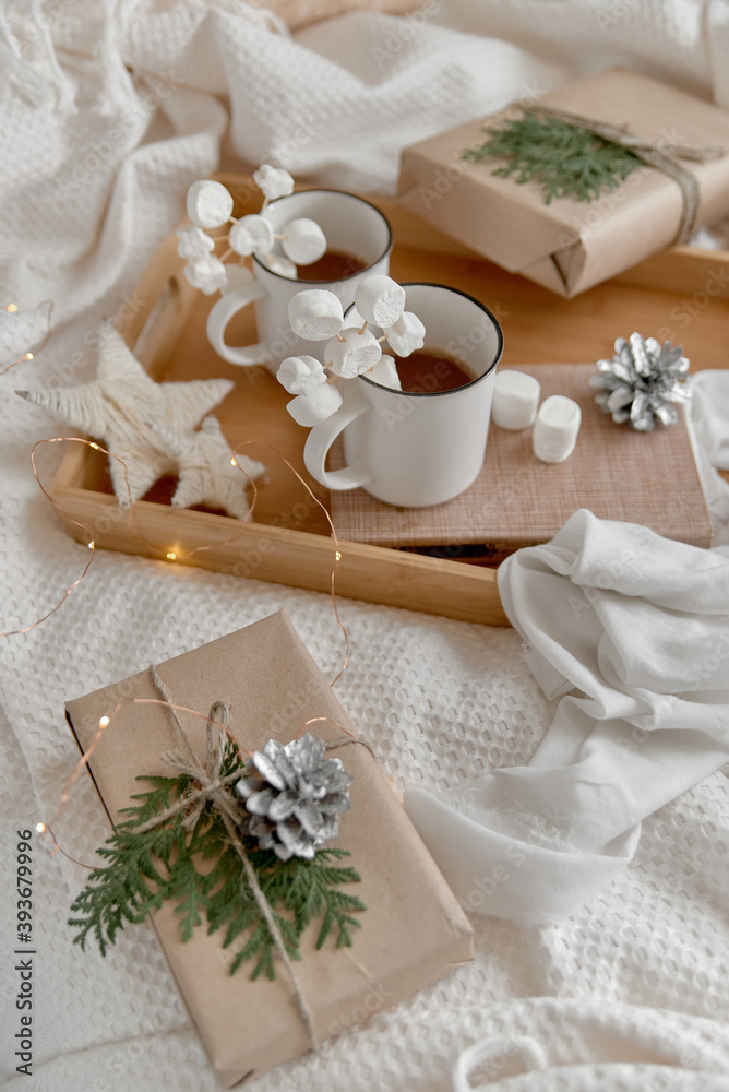 Naklejka premium Hot cocoa with marshmallow in a white ceramic mug, on a wooden tray and wrapped boxes. The concept of cosy holidays and New Year. Selective focus