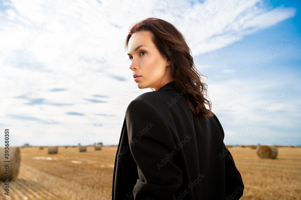 Fototapeta premium a young beautiful girl in a black suit in summer on a hot day stands in a field with haystacks