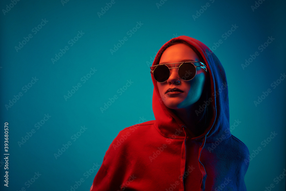 Young model posing in a studio in a trendy neon light. Woman in round sunglasses and hoodie with colorful light effects.