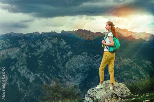  portrait of a modern woman tourist with a backpack standing on a rosk and looking around of landscaping views on background of the mountain range, cloudy sky and sun lights.
